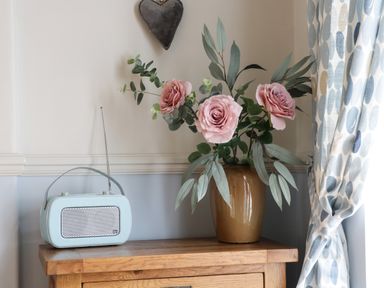 A wooden table with a retro radio and a vase with artificial pink roses and green leaves next to patterned curtains at Potter Hill Cottage in Pickering
