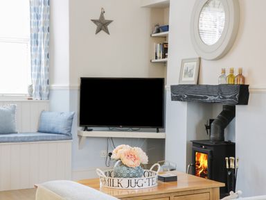 A living room with a wall-mounted TV window seat cushions a fireplace with a wood stove and a coffee table with flowers at Potter Hill Cottage in Pickering