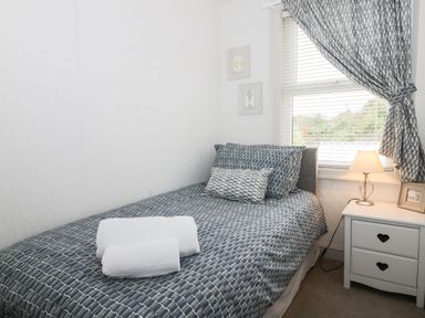 A bedroom with a single bed with patterned bedding two rolled towels on the bed a window with blinds and matching curtains and a bedside table with a lamp and picture frame at Potter Hill Cottage in Pickering