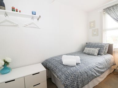 A small bedroom with a single bed covered with patterned bedding and two rolled towels a white dresser with flowers and a wall shelf with hangers at Potter Hill Cottage in Pickering
