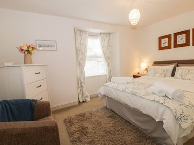 A bedroom with a bed with floral bedding and rolled towels a white dresser with a vase and flowers a brown armchair a window with floral curtains and framed pictures on the wall at Potter Hill Cottage in Pickering