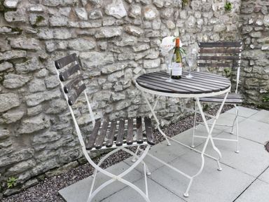 A small outdoor seating area with two wooden slat chairs and a round wooden slat table with a wine bottle and glasses against a stone wall at Potter Hill Cottage in Pickering