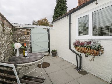 An outdoor patio with a wooden table and chairs a bottle of wine glasses a flower vase stone wall a white gate and a flower planter on the white brick wall at Potter Hill Cottage in Pickering