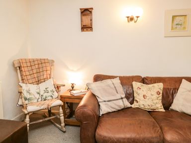 A corner of a living room with a leather sofa with cushions a wooden rocking chair with cushions and a blanket a side table with a lamp and a book on it at Beckfold Cottage in Broughton-In-Furness