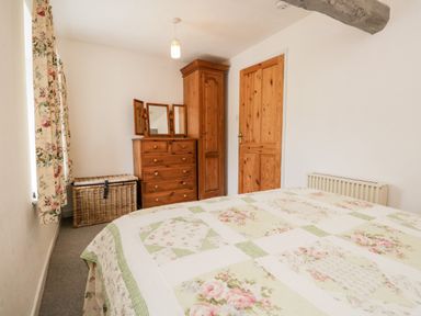 A bedroom with a bed covered by floral patterned quilt a wooden dresser with mirror a wicker basket and a wooden wardrobe at Beckfold Cottage in Broughton-In-Furness