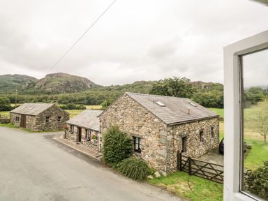 A stone cottage and outbuilding next to a road with mountains and trees in the background at Beckfold Cottage in Broughton In Furness