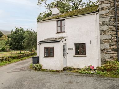 A white cottage with brown windows and a sign reading Beckfold Cottage next to a road with trees and plants at Beckfold Cottage in Broughton-In-Furness
