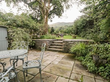 An outdoor stone patio with metal chairs and table a stone step leading to a grassy area with a dog and picnic bench at Beckfold Cottage in Broughton-In-Furness