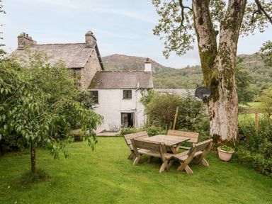 A garden with wooden table and benches under a tree behind a stone and white cottage at Beckfold Cottage in Broughton-In-Furness