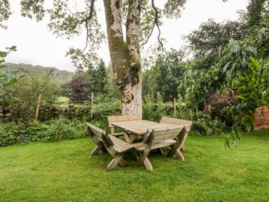 A wooden picnic table with benches under a tree in a garden with green grass and plants at Beckfold Cottage in Broughton-In-Furness