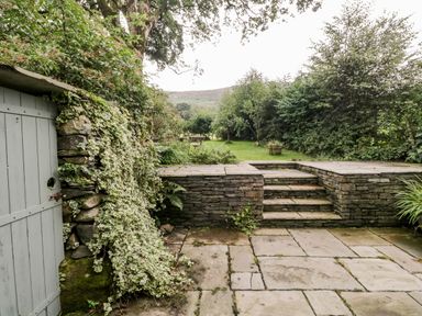 An outdoor stone patio with steps leading to a grassy garden with trees and plants at Beckfold Cottage in Broughton-In-Furness
