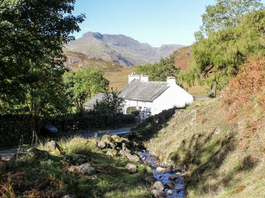 A white cottage near a small stream with trees and mountains in the background at Beckfold Cottage in Broughton-In-Furness