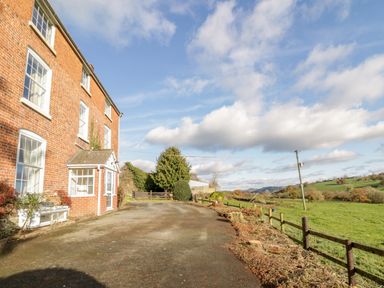 A brick house with multiple windows and a small porch next to an open paved area and fenced green fields at Squires Rest in Llanfair Caereinion