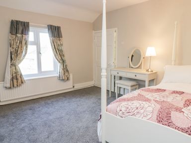 A bedroom with a bed with floral bedding a dressing table with a mirror and lamp and a window with curtains at Squires Rest in Llanfair Caereinion