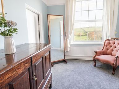 A room with a wooden cabinet with a vase of flowers a full length mirror a window with curtains and a pink upholstered armchair at Squires Rest in Llanfair Caereinion