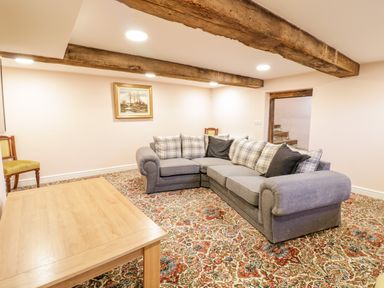 A room with a large gray sectional sofa with cushions a wooden table two chairs a framed painting on the wall exposed wooden ceiling beams and a patterned carpet at Squires Rest in Llanfair Caereinion