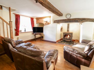 A living room with a wood burning stove two brown leather sofas a television red curtains and wooden beams at Squires Rest in Llanfair Caereinion