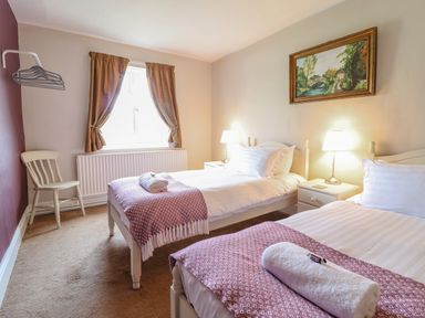 A bedroom with two single beds side by side each with a pillow blanket and rolled towel a chair under a window with curtains two bedside tables with lamps and a framed picture on the wall at Squires Rest in Llanfair Caereinion