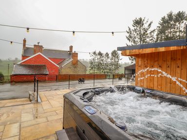 An outdoor area with a bubbling hot tub a wooden shed stone pavement and string lights with red brick buildings and trees in the background at Squires Rest in Llanfair Caereinion