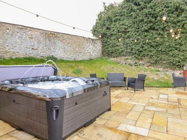 An outdoor patio with a hot tub and wicker chairs near a stone wall and greenery at Squires Rest in Llanfair Caereinion