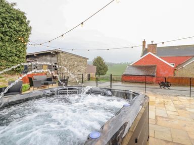 An outdoor hot tub with water jets bubbling and string lights above with a stone building and red house in the background at Squires Rest in Llanfair Caereinion