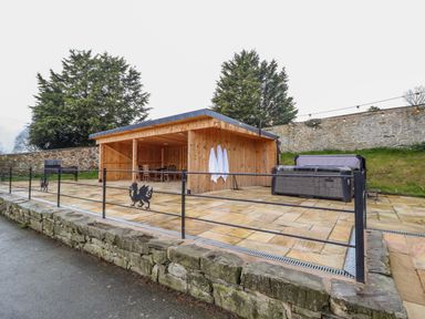 An outdoor wooden shelter with chairs and table next to a hot tub and grill on a stone patio at Squires Rest in Llanfair Caereinion