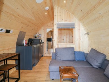 A small wooden pod interior with a kitchenette a grey sofa a wooden table and a doorway with a TV visible through it at Garraidh Ghorm Pod in Conista