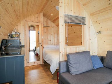 A small wooden room with a bed visible through a glass door a sofa with cushions and a kitchenette area at Garraidh Ghorm Pod in Conista