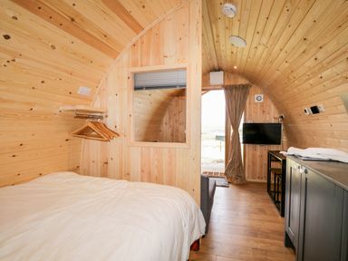 A wooden interior pod with a bed a window with a blind a hanging rack with hangers a mounted television and a kitchenette at Garraidh Ghorm Pod in Conista