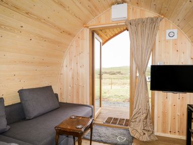 A small wooden pod interior with a gray couch a small wooden side table a wall-mounted television and a door opening to a grassy outdoor area at Garraidh Ghorm Pod in Conista