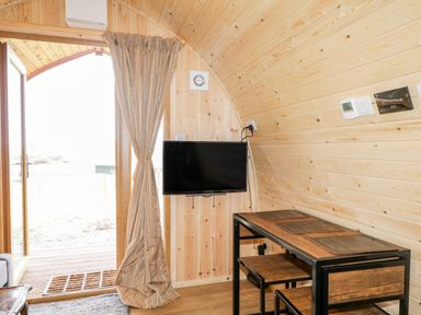 A wooden room with a small table and chairs a wall-mounted TV and a door with curtains at Garraidh Ghorm Pod in Conista