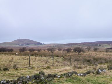 A rural landscape with grass fields and sparse trees with hills and houses in the distance at Garraidh Ghorm Pod in Conista