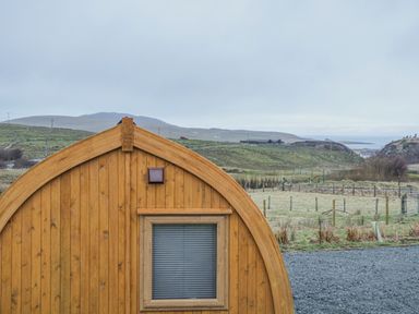 A curved wooden cabin with a window and light in a rural landscape at Garraidh Ghorm Pod in Conista