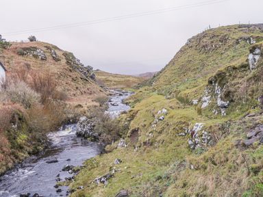 A river flowing through a grassy valley with rocky hills on both sides at Garraidh Ghorm Pod in Conista
