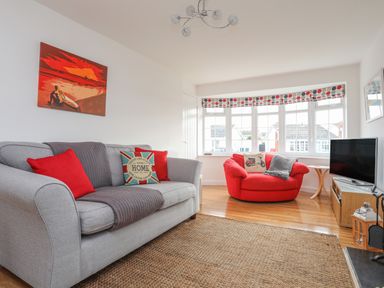 A living room with a gray sofa with red and patterned cushions a red armchair a wall painting window with patterned blinds and a television on a wooden stand at Sisial-Y-Mor in Rhosneigr