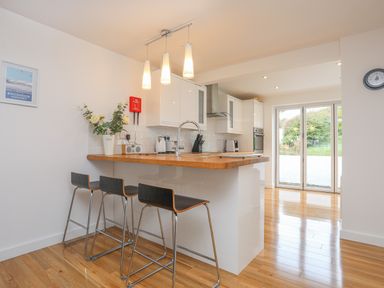 A kitchen with a wooden countertop bar with three stools white cabinets and glass doors at Sisial-Y-Mor in Rhosneigr