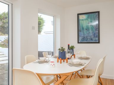 A dining area with a white oval table set with plates bowls wine glasses and cutlery four beige chairs two potted plants and a wine bottle on the table a poster on the wall a glass door and window showing an outdoor area at Sisial-Y-Mor in Rhosneigr