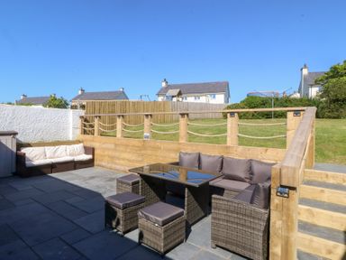 An outdoor patio with wicker furniture including a sofa and stools a table wooden railing and steps leading to a grassy area with houses in the background at Sisial-Y-Mor in Rhosneigr