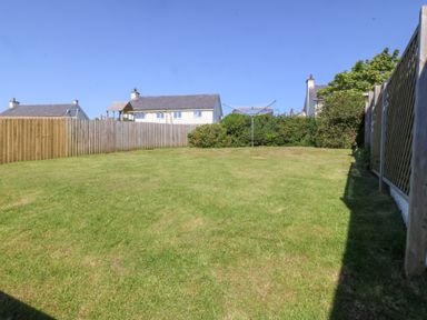 A fenced backyard with grass a clothesline and houses in the background at Sisial-Y-Mor in Rhosneigr