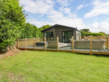 A wooden house with glass doors and outdoor seating on a fenced deck surrounded by grass and trees at Pinehurst Lodge in St Day