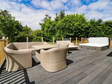 An outdoor deck with wicker chairs a table and a hot tub surrounded by greenery at Pinehurst Lodge in St Day