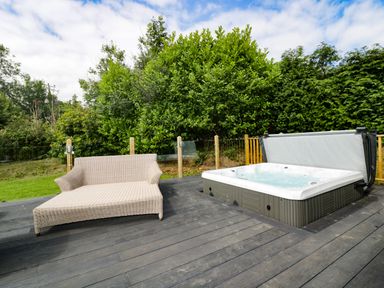 An outdoor wooden deck with a wicker sofa and a built-in hot tub surrounded by greenery at Pinehurst Lodge in St Day