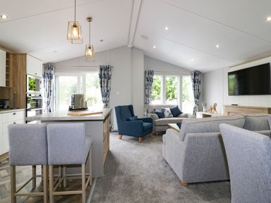 A living room with grey sofas a blue armchair a kitchen island with bar stools and a mounted television at Pinehurst Lodge in St Day