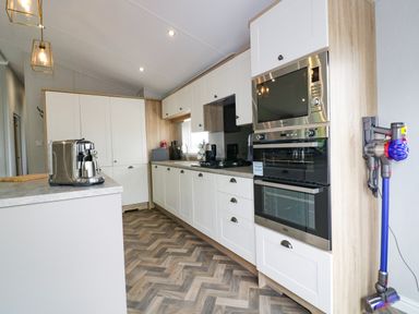 A kitchen with white cabinets a coffee machine built-in oven microwave and a vacuum cleaner on the wall at Pinehurst Lodge in St Day