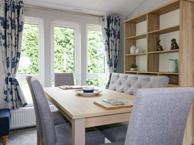 A dining area with a wooden table surrounded by gray chairs and a bench near windows with floral curtains and a wooden shelving unit at Pinehurst Lodge in St Day