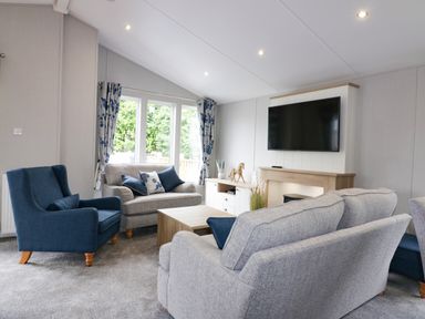 A living room with a gray sofa set a blue armchair a wooden coffee table a wall-mounted television and windows with curtains at Pinehurst Lodge in St Day