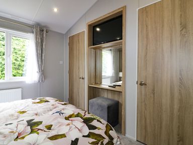 A bedroom with a floral bedspread a window with curtains a built in vanity with a mirror a stool and a wall mounted TV at Pinehurst Lodge in St Day