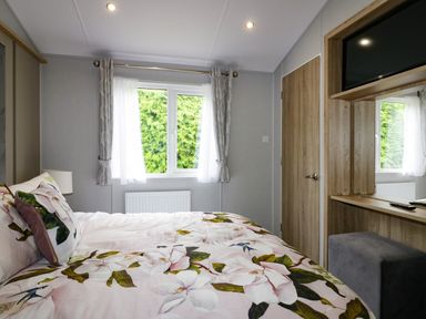 A bedroom with a bed covered in floral bedding a window with curtains a lamp on a bedside table and a wooden wardrobe with a mounted tv at Pinehurst Lodge in St Day