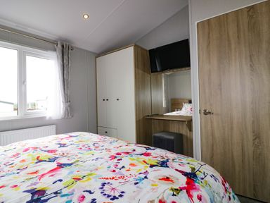 A bedroom with a bed featuring a colorful floral bedspread a wardrobe a wall-mounted television a mirror and a window with curtains at Pinehurst Lodge in St Day