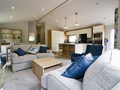 An open plan living room and kitchen with grey sofas blue cushions wooden coffee table and white cabinets at Pinehurst Lodge in St Day
